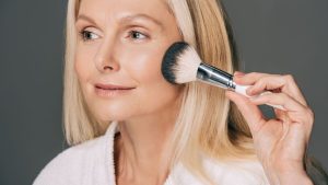 Женщина наносит лёгкую жидкую тональную основу на щёку, естественный макияж для зрелой кожи, мягкий свет и ухоженное лицо