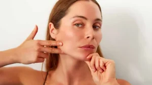 Женщина выполняет упражнения йоги для лица перед зеркалом; массаж лица для повышения тонуса кожи; упражнения против морщин и второго подбородка.