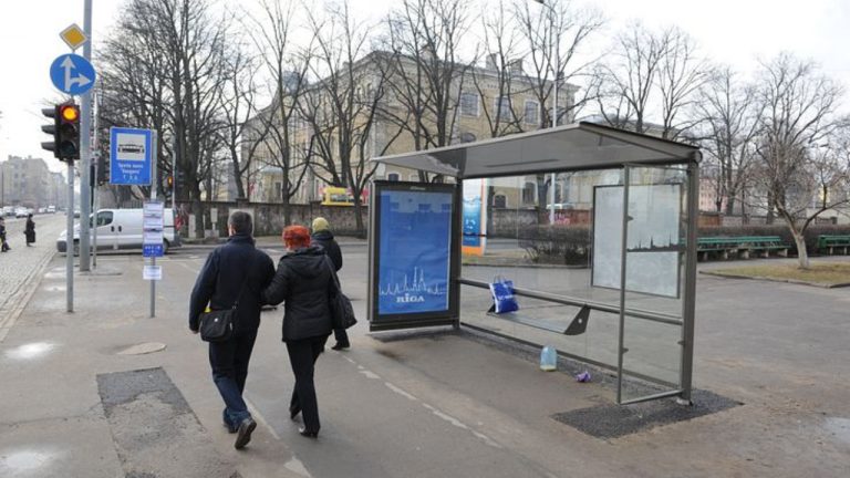 Новый павильон остановки общественного транспорта в Риге с современным стеклянным навесом и городской улицей на фоне.