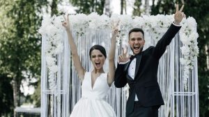 Счастливая пара молодожёнов на свадьбе: невеста в белом платье с букетом и жених в костюме, праздничная атмосфера и гости на фоне.